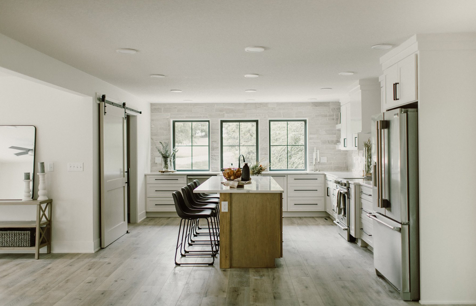 Whole kitchen view with refrigeration on right, three large windows behind an island and barn door on the left