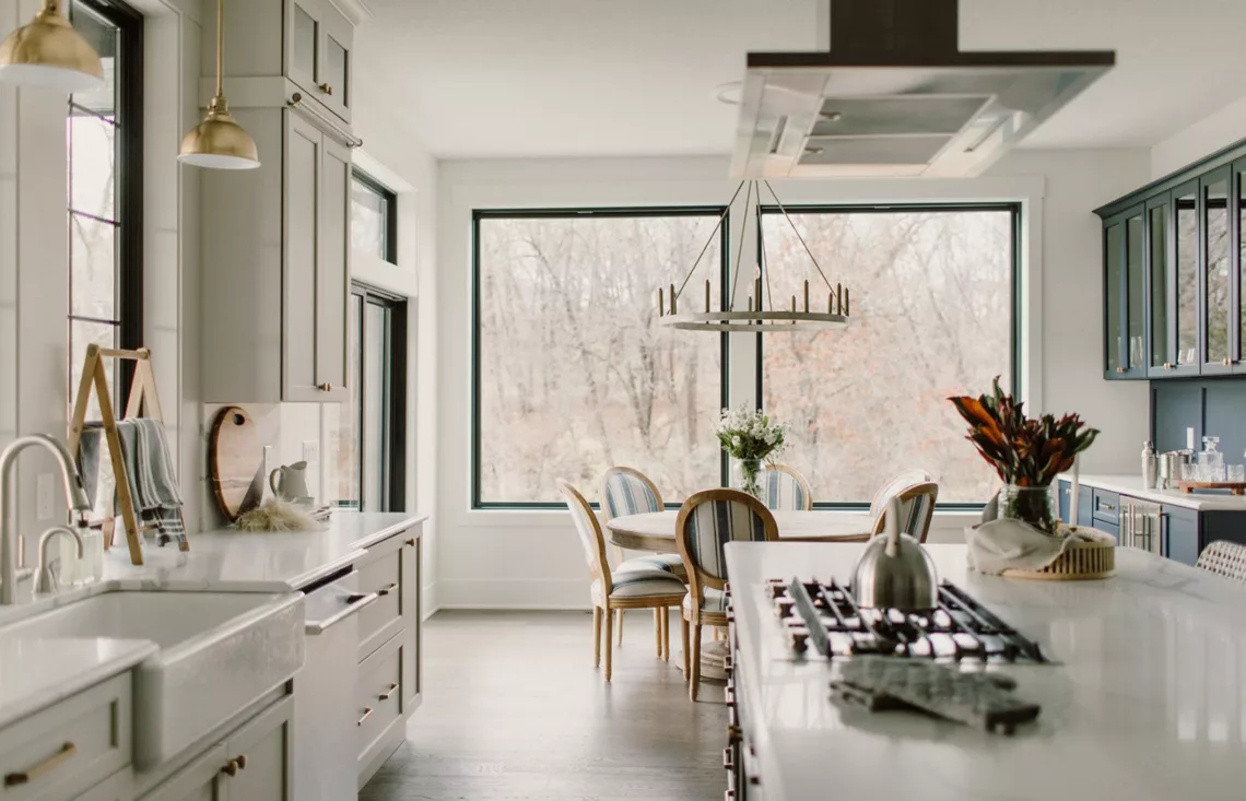 large windows behind dining area and large open kitchen with white counters and light colored cabinets with gold hardware