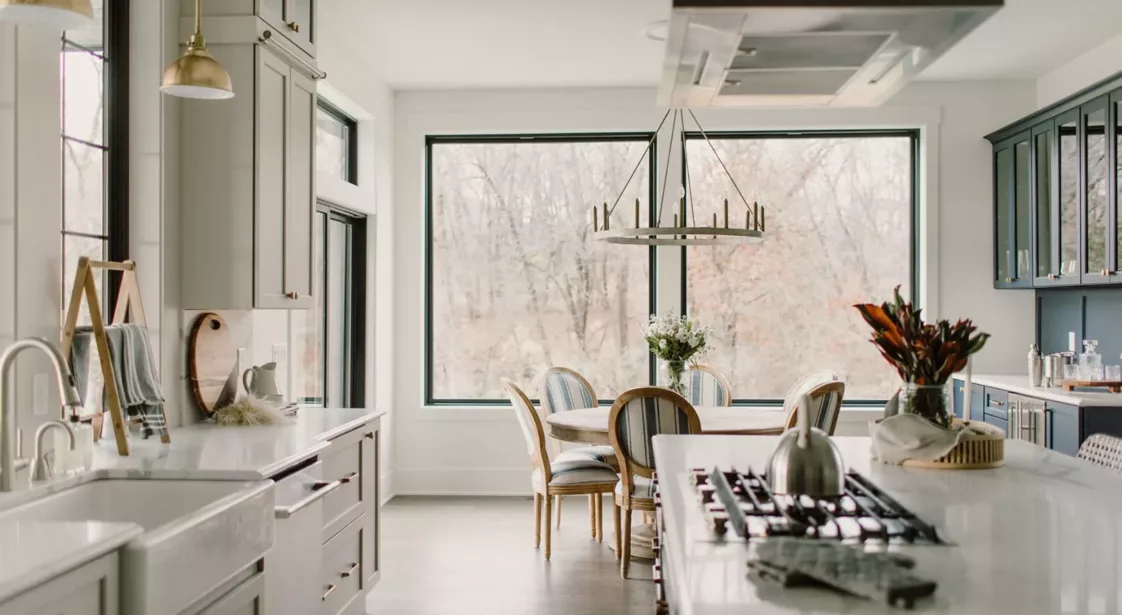 large windows behind dining area and large open kitchen with white counters and light colored cabinets with gold hardware