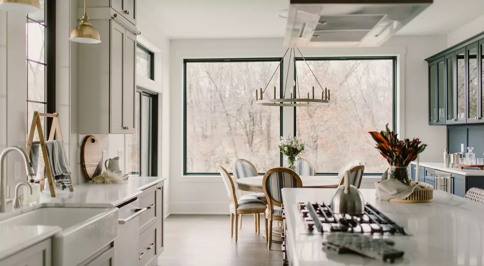 large windows behind dining area and large open kitchen with white counters and light colored cabinets with gold hardware