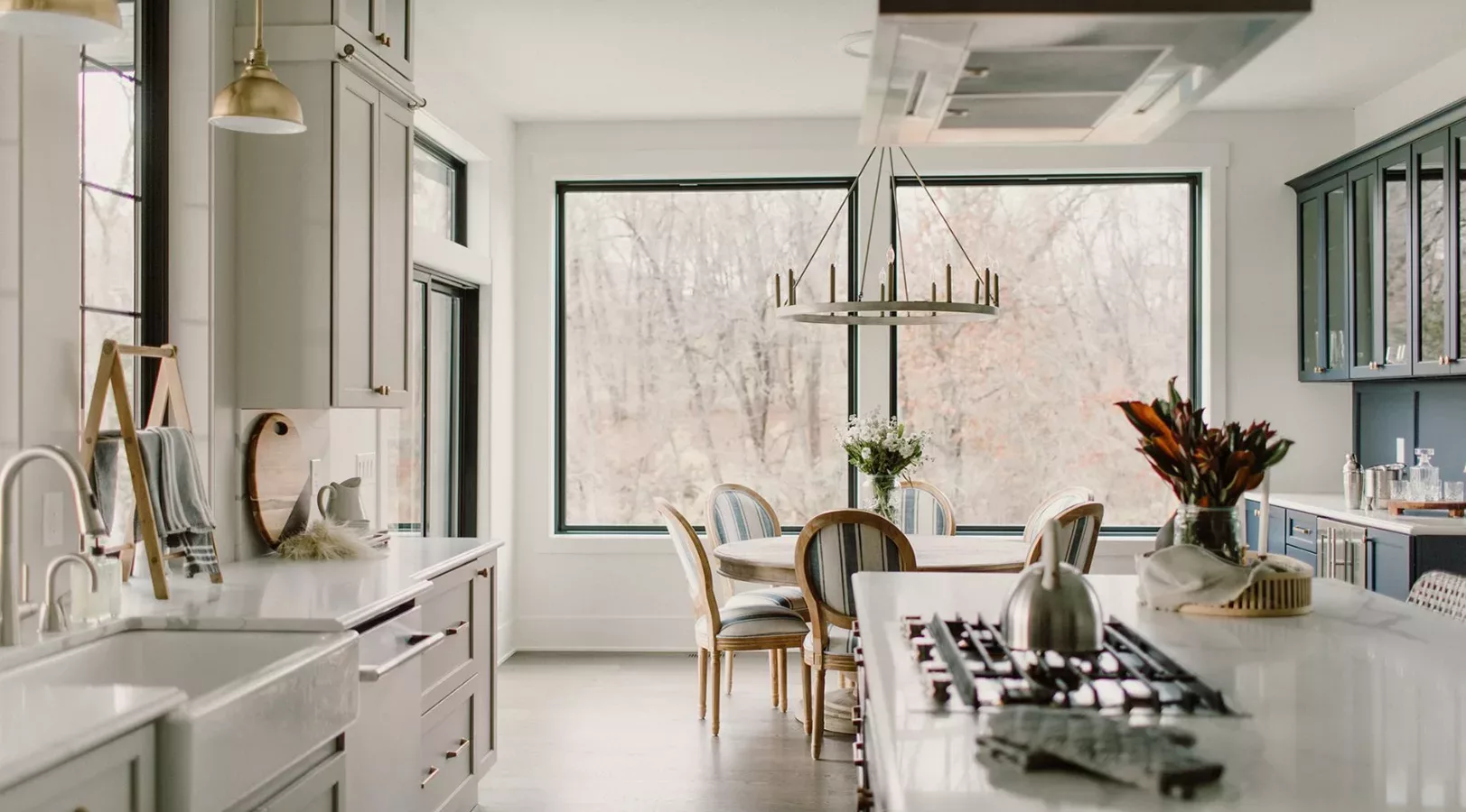 large windows behind dining area and large open kitchen with white counters and light colored cabinets with gold hardware