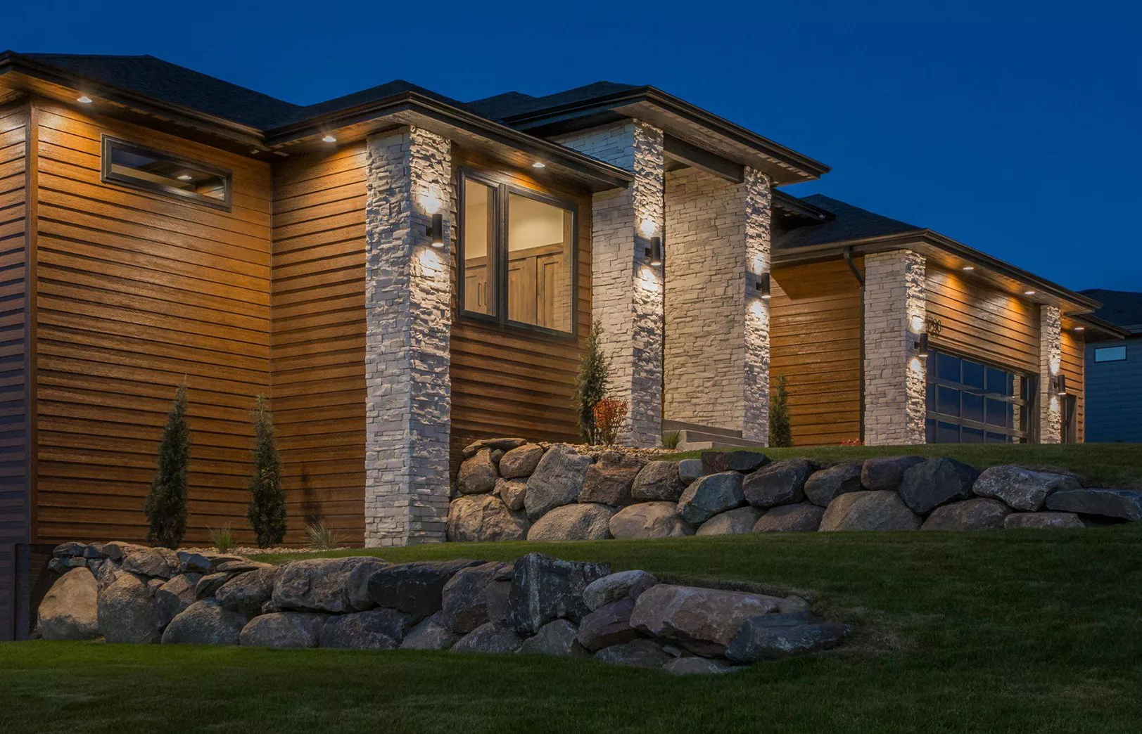 lighted home exterior with stained wood siding and stacked stone pillars