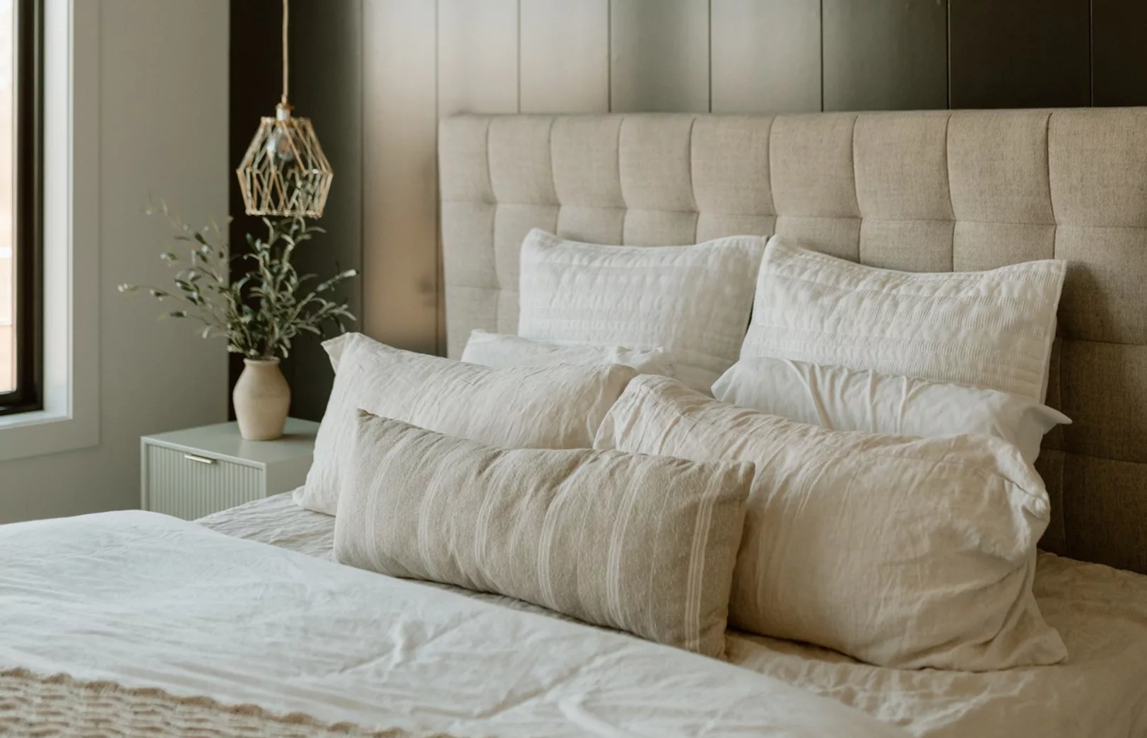 black accent wall with gold wire lighting fixture over light sage green side table next to bed with cream colored linens and pillows